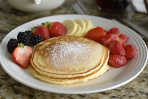A plate of fluffy golden pancakes dusted with powdered sugar, surrounded by fresh sliced strawberries, blackberries, and banana slices, presented on a white plate with a warm, inviting table setting.