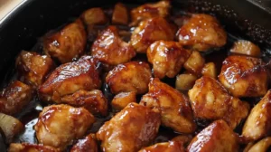 Close-up of golden-brown glazed chicken bites cooked with caramelized onions and a rich sauce in a skillet, glistening with flavor.